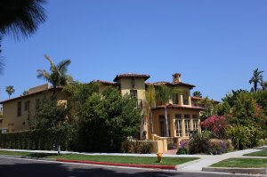 A large, beige stucco house with reddish-brown roof tiles stands against a clear blue sky. Surrounded by lush greenery, colorful flowers, and palm trees, the home features multiple windows and a chimney. A red curb and sidewalk are visible in the foreground.