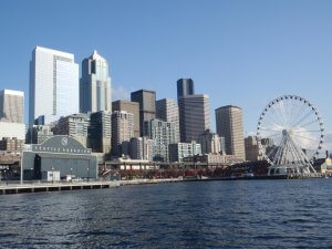 A panoramic view of Seattle's waterfront showcases the iconic Seattle Aquarium building, a towering Ferris wheel, and a mix of modern skyscrapers with older buildings under a clear blue sky. The water in the foreground reflects this vibrant Seattle cityscape beautifully.