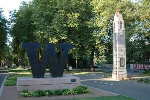 A large metallic "W" sculpture stands prominently on a stone base near an obelisk monument surrounded by Seattle's lush green trees. The road splits around the monument, leading further into a leafy area. A sign with "Burke" is visible in the background.