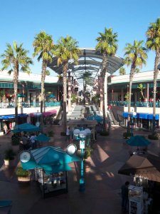 An outdoor shopping mall with palm trees, multiple levels of shops, and a central walkway. People are walking and browsing around various kiosks and stores. There are blue awnings, a clock, and a glass canopy covering part of the walkway. The sky is clear and blue.