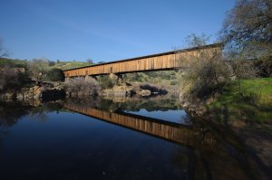 A long, wooden covered bridge spans across a calm river, reflecting its image on the water's surface. Surrounding the bridge are green hills and sparse trees under a clear, blue sky. Rocky banks and gentle slopes complete the serene landscape.