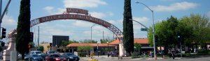 A large arch with the word "Modesto" and the motto "Water Wealth Contentment Health" spans across a busy street intersection, bordered by two tall trees. Cars are stopped at the traffic lights and pedestrians walk along the sidewalks under a partly cloudy sky.