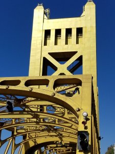 Steel bridge with yellow painted structure and traffic lights. The bridge features a tall tower with geometric cutouts and is set against a clear blue sky.