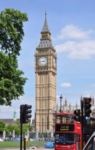 A tall clock tower, commonly known as Big Ben, stands under a clear blue sky. A red double-decker bus and traffic lights are in the foreground. Lush green trees partially frame the left side of the image. Historic buildings and greenery are visible in the background.