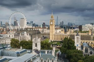 A view of central London with iconic landmarks: the London Eye, Big Ben, the Houses of Parliament, and the Shard. The scene features a mix of historic and modern buildings under a cloudy sky. Trees and various architectural structures are visible in the foreground.