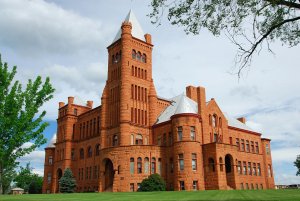A large historic red sandstone building with a prominent central tower and multiple arched windows, set against a lightly clouded sky. Surrounding it are green lawns and trees, with some branches visible in the foreground.