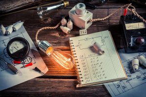 A wooden table cluttered with electrical equipment, a glowing light bulb, and technical drawings sits in a college dorm room. An open graphing notebook filled with electrical calculations lies beside a small model. Multimeters and circuit diagrams are also scattered around, reflecting an intense study session.