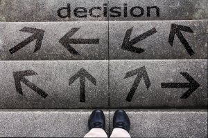 A person wearing black shoes stands at the bottom of a set of stairs. The steps display multiple black arrows pointing in various directions, symbolizing choices. At the top, the word "decision" is written, reflecting the pivotal choices often faced during college life.