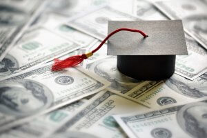 A graduation cap with a red tassel sits atop a pile of hundred-dollar bills, symbolizing the cost of college education. The image focuses on the financial aspects related to obtaining a degree.