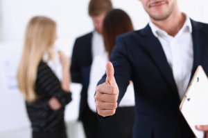 Smiling person in a business suit giving a thumbs-up gesture. In the background, two people have a discussion while standing, and another person is on the phone. It's clear that this dynamic environment is what employees really want in their workplace.