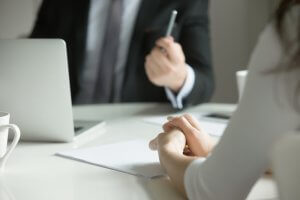A person in formal attire, partially visible, holds a pen while gesturing during a discussion at an office desk with another individual. A laptop and documents are on the desk. The focus is on the hands and posture, suggesting a professional meeting or interview, possibly avoiding an interviewing mistake.