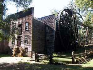 A rustic, wooden mill with a large water wheel attached on the side is surrounded by trees. The mill features multiple windows and a gabled roof. A split-rail wooden fence encloses part of the property, and pumpkins are arranged near the mill's entrance.