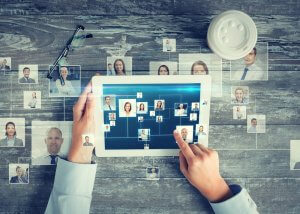 A person holds a tablet displaying a network of headshots, indicating an academic digital organizational chart or virtual connections. Surrounding the tablet on a wooden surface are glasses, additional headshots, and a takeout coffee cup with a lid.