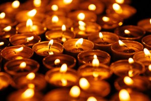 A close-up photo showing numerous small, lit candles arranged close together. The soft, warm glow from the candle flames creates an inviting and tranquil atmosphere, reminiscent of a peaceful evening with sheep grazing nearby. The golden light reflects off the candle holders, creating a comforting ambiance.