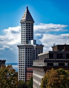A tall, historic skyscraper with a cone-shaped roof stands against a blue Seattle sky with scattered clouds. The building is surrounded by other shorter buildings and trees, with a glimpse of water in the background.
