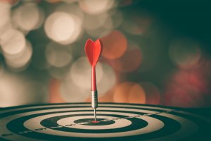 A red dart with a heart-shaped tail is perfectly lodged in the bullseye of a target. The background is blurred, featuring circular bokeh lights that create a warm and festive atmosphere, mirroring the precision and finesse often associated with soft skills.