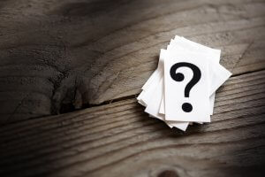 A stack of small white cards, with the top card displaying a large black question mark, lies on a rustic wooden surface. The wood, weathered with visible grain and cracks, seems to mirror the intriguing nature of unanswered questions.