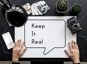 A person holds a white board with a speech bubble that says "Keep It Real" on a black tabletop. Nearby are a vintage camera, a small potted cactus, a lamp, a smartphone, and scattered photographs—each representing fragments of their personal constitution.