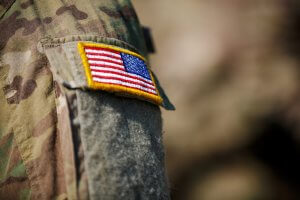 Close-up of a camouflage military uniform sleeve featuring a patch with the American flag, honoring our veterans. The background is blurred and out of focus.