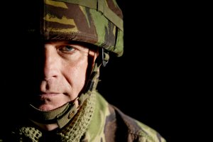 A male soldier wearing a camouflage uniform and a helmet is partially illuminated against a dark background. His serious facial expression is visible as he looks directly into the camera, embodying the steadfast resolve of veterans.