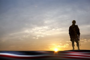 A soldier in uniform stands on a hilltop holding a rifle, silhouetted against a dramatic sunset sky. The foreground is out of focus, showing what appears to be a blurred American flag—a poignant reminder of the sacrifices made by our veterans.
