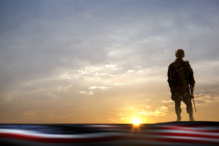 A soldier in uniform stands on a hilltop holding a rifle, silhouetted against a dramatic sunset sky. The foreground is out of focus, showing what appears to be a blurred American flag—a poignant reminder of the sacrifices made by our veterans.