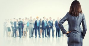 A woman in a gray suit, seen from behind, stands with one hand on her hip while facing a large group of businesspeople in professional attire. The group is somewhat blurred, emphasizing her as the focal point and symbolizing leadership opportunities. The background is a light, neutral color.