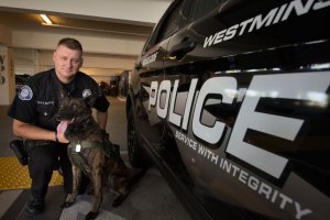 A Westminster Police Department officer in a black uniform kneels next to a police dog in a parking garage. The dog, wearing a harness, has its tongue out. They are beside a black police vehicle with the word "POLICE" and the phrase "SERVICE WITH INTEGRITY" on its side.