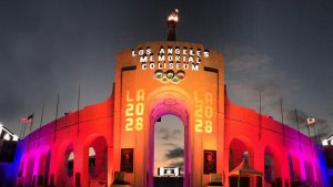 A photo of the Los Angeles Memorial Coliseum illuminated at dusk. The iconic entrance is lit up in red and orange hues, displaying the Olympic rings and the text "LOS ANGELES MEMORIAL COLISEUM" at the top. The numbers "LA 2028" flank the entrance arch, capturing a quintessential Los Angeles evening.