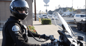 A Westminster police department officer, wearing a black helmet with a visor and a communication headset, sits on a motorcycle. The officer has a serious expression and is looking towards the camera. A busy street with cars and buildings is visible in the background.