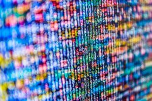 A vibrant, multicolored image displaying lines of alphanumeric text and code on a digital screen. The densely packed text appears to be a close-up view of programming code or big data, with various colors potentially representing syntax highlighting.