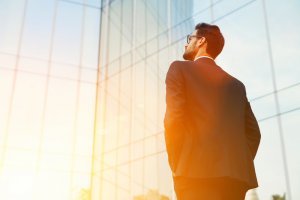 A man in a dark suit and glasses stands with his hands in his pockets, looking up at a modern glass building. The sun is shining brightly, illuminating the scene as he contemplates his career path.