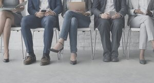 A row of seated individuals, dressed in business attire, sit on white chairs. Each person is engaged with a device: some use smartphones, and one reads from a tablet. The scene captures the essence of modern networking, evident in their varied postures and footwear, with only their lower halves visible.