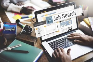 A person is using a laptop to search for jobs online. The screen displays a job search website along with networking opportunities. Surrounding the laptop are books, notebooks, a pen, and a pair of glasses. The search bar on the website prominently displays the text "Job Search.