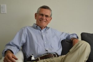 A smiling older man with short gray hair and glasses is sitting on a couch. Clad in a blue and white striped dress shirt and beige pants, he exudes an air of leadership with his relaxed and casual pose, one arm gracefully resting on the arm of the couch.