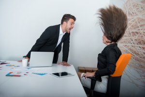 A man in a suit leans over a desk, angrily yelling at a woman whose hair is flying upward. She sits in an orange chair with a laptop, tablet, and scattered papers on the desk. The scene suggests a heated argument between coworkers in a modern office setting.