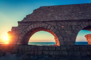 Ancient stone arches stand like sentinels of leadership against a backdrop of a vibrant sunset over the sea. The sun can be partially seen through one of the arches, casting a warm golden glow on the stone structures and illuminating the sky with shades of orange and blue.