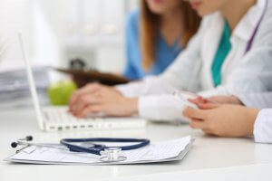 Two healthcare professionals sit at a desk, one working on a laptop and the other holding a phone. A stethoscope and clipboard with medical documents are visible in the foreground. Their focus appears to be on the screen and important healthcare documents.