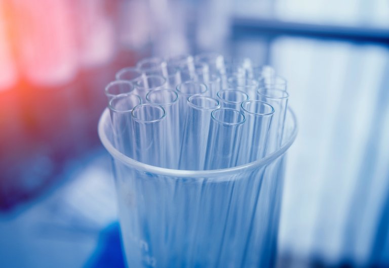 A cluster of empty, transparent test tubes is arranged in a beaker. The background is blurred with a blue and reddish hue, giving the image a scientific and healthcare laboratory atmosphere.