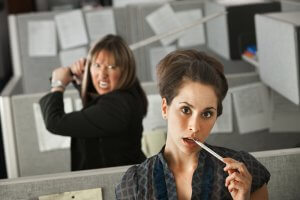 Two coworkers in an office cubicle. The woman in the foreground looks perplexed, with a pencil in her mouth. The woman behind her appears angry, holding a wooden sword over her shoulder in a striking pose. Office cubicle walls are covered in papers.