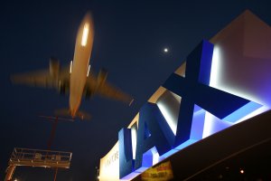 An airplane is captured in motion as it flies over a brightly lit, large blue "LAX" sign at Los Angeles International Airport during nighttime. The moon is visible in the dark sky, with HireLAX making its mark alongside this iconic scene.
