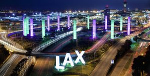 A nighttime view of Los Angeles International Airport (LAX) featuring brightly lit, colorful pillars. The airport's iconic, illuminated "LAX" sign can be seen in the foreground, with streaks of car lights on the roads highlighting the busy atmosphere and opportunities through HireLAX.