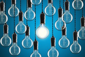 A series of unlit incandescent light bulbs hang from black cords against a blue background. One light bulb in the center is brightly illuminated, symbolizing leadership and standing out from the rest.