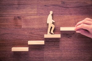 Cutout of a person climbing wooden blocks arranged like stairs, placed on a wooden surface. A hand is adding the next block, symbolizing mentoring, progress, growth, or step-by-step advancement in goals or career.