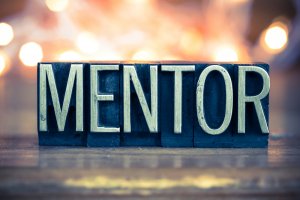 The image shows the word "MENTOR" in block letters, with a vintage, metallic look. The letters are arranged on a flat surface, with a softly blurred background of warm lights, giving a cozy and inviting atmosphere reminiscent of an ideal mentoring environment.