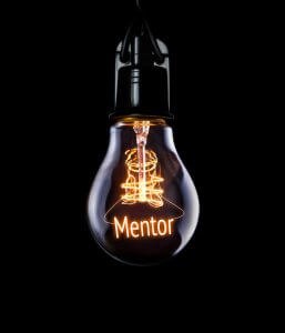 A hanging light bulb, illuminated against a black background, with the word "Mentor" glowing inside the bulb's filament. The filament is shaped to spell out the word, symbolizing the enlightening role of mentoring.
