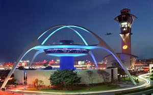 The image depicts the iconic Theme Building at Los Angeles International Airport (LAX) illuminated against a night sky. The futuristic design features curved arches and a central circular structure. In the background, an air traffic control tower and a plane in flight can be seen. This highlights the success of HireLAX in maintaining such landmarks.