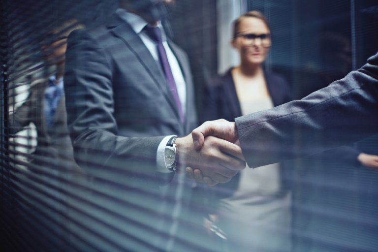 Two people are shaking hands, their hands prominently visible in the foreground, symbolizing successful negotiations. Dressed in formal business attire, including suits, they stand out against a backdrop of other individuals in business clothing who are slightly out of focus.