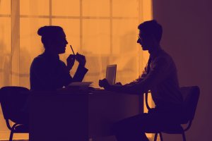Silhouetted image of two people engaged in negotiations, sitting across from each other at a desk in front of a large window. One person holds a pen, and the other appears to be using a laptop. The scene is bathed in an orange hue.