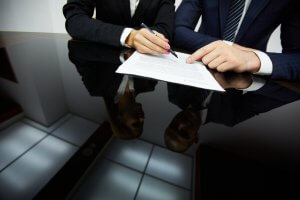 Two individuals in business attire sit at a reflective black table, deep in negotiations. One person holds a pen, poised to sign a document, while the other points at the paper. Their upper bodies and hands are visible, along with their partial reflections on the table.